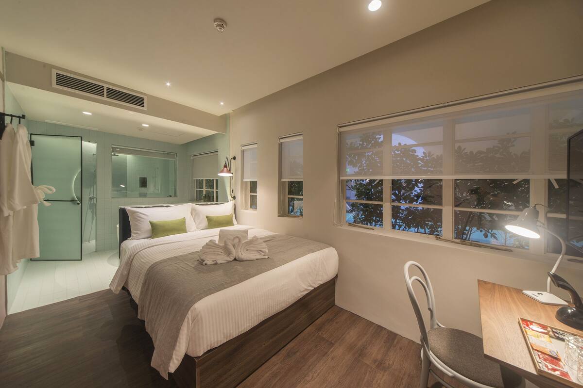 Une chambre moderne avec un grand lit, un bureau avec une lampe et une vue sur les arbres par la fenêtre.