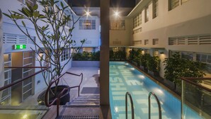 Una piscina con un cartel que dice 'WHILE SHINING', rodeada de edificios y plantas en macetas.