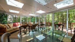 A sunlit room with a glass table surrounded by chairs, large windows, and a view of a wooded area.