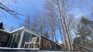A hot tub with a wooden deck, surrounded by trees and a house with large windows.