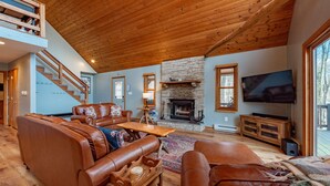 A spacious living room with a stone fireplace, leather sofas, a wooden coffee table, and a TV mounted on the wall. - Rustic Woodsy Getaway w/Hot Tub & Fireplace (Oakland)