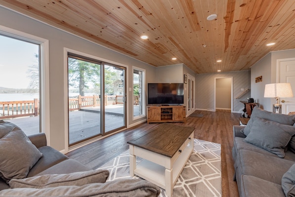 A spacious living room with a wooden ceiling, large sliding glass doors leading to an outdoor deck, a flat-screen TV, and a cozy seating area with a sofa and a coffee table.