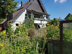 Ein zweistöckiges Haus mit einem steilen Dach, einem Balkon und einem Garten mit verschiedenen Pflanzen und Blumen.