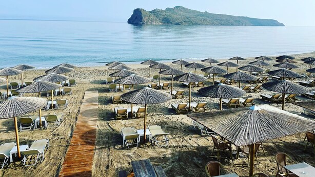 Una playa con hileras de tumbonas y sombrillas de paja, un paseo de madera, y vistas al mar y una isla distante.