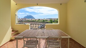 En balkon med et træbord og stole, udsigt til bygninger og bjerge, samt et flisegulv.