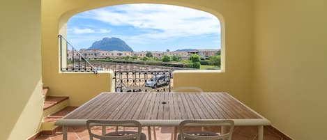 A balcony with a wooden table and chairs, a view of buildings and mountains, and a tiled floor.