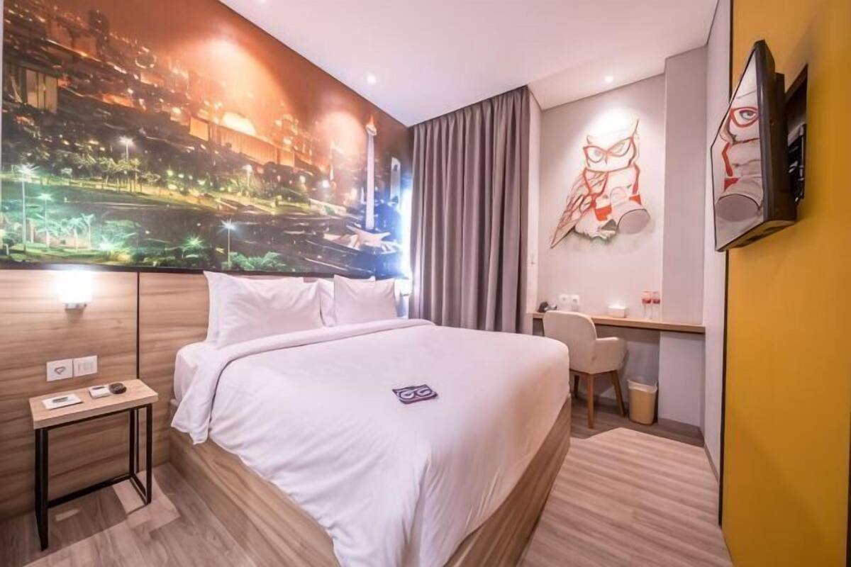 A hotel room with a bed, a desk, a chair, a wall-mounted TV, and a large cityscape mural.