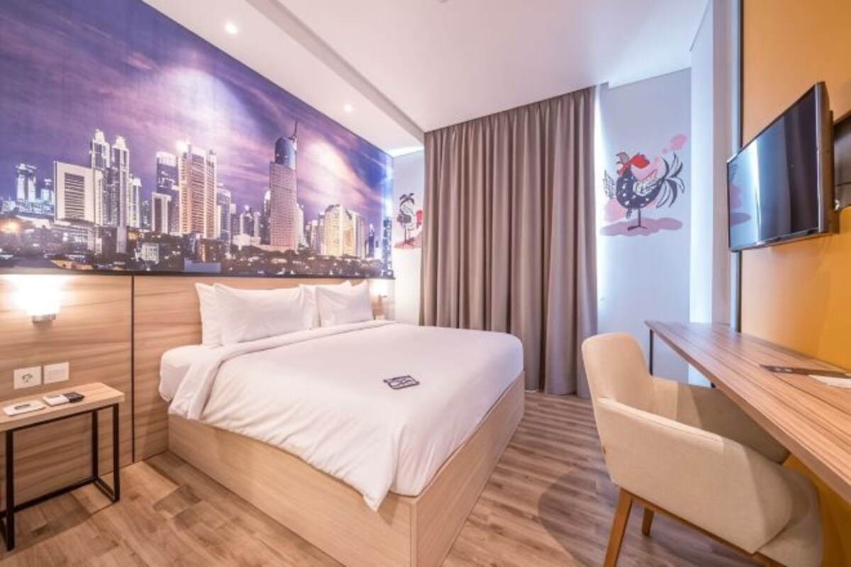 A hotel room with a large bed, a desk, a chair, a television, and a cityscape mural on the wall.