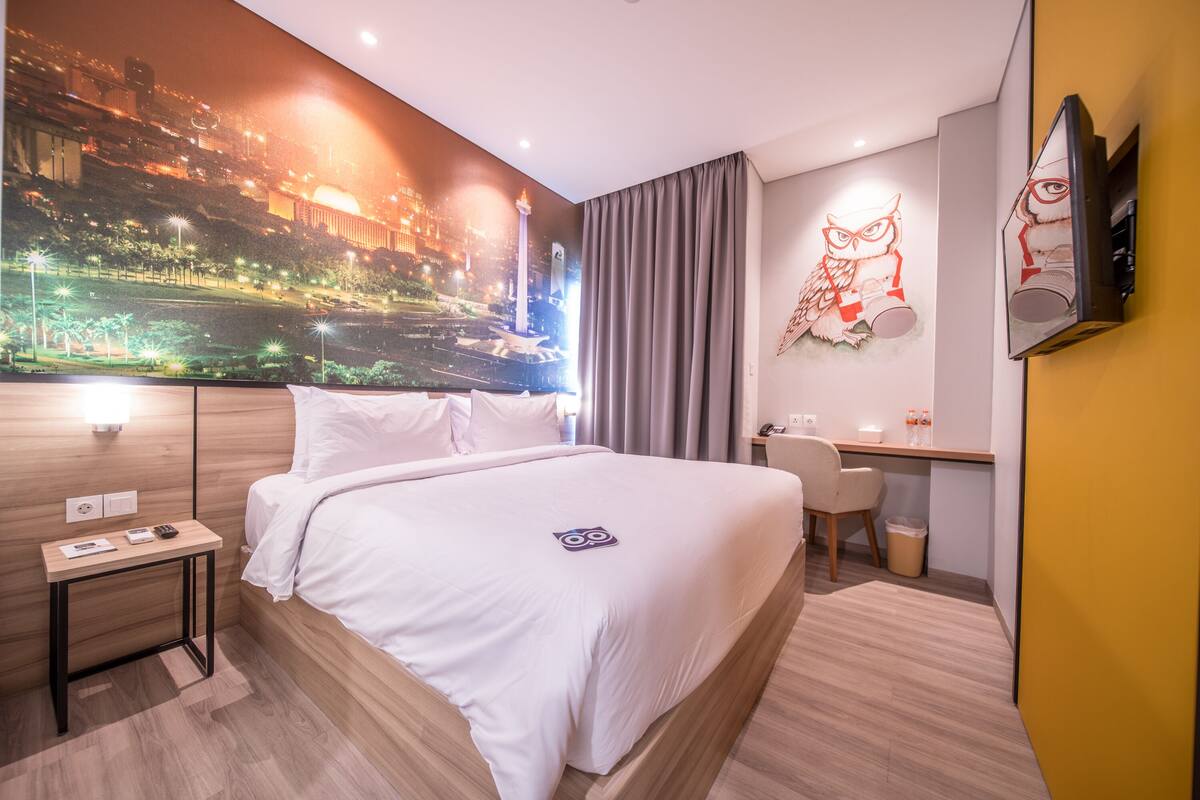 A hotel room with a large bed, a desk, a chair, a TV, and a mural on the wall.