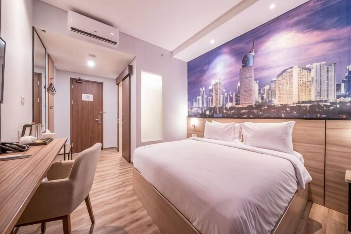 A hotel room with a bed, a desk, a chair, a cityscape painting, and an air conditioning unit.