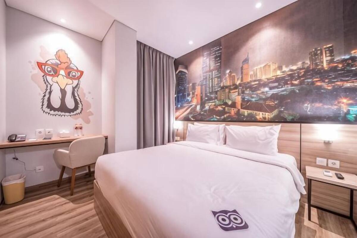 A hotel room with a bed, a desk, a chair, a wall with a cityscape painting, and a wall-mounted owl decoration.