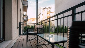 A balcony with a glass table and a cushioned chair, overlooking a garden and adjacent buildings.
