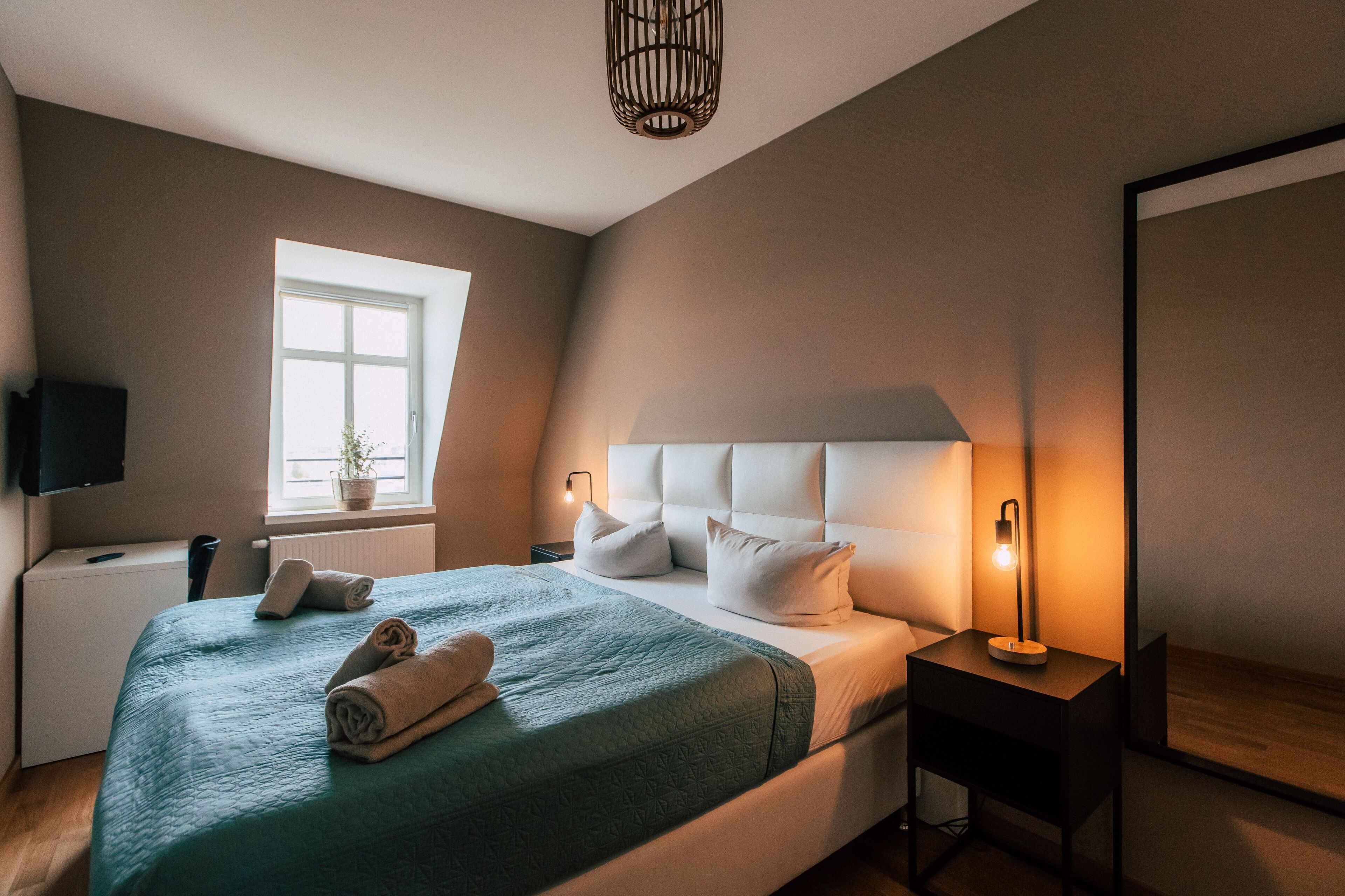 Una cama bien tendida con una colcha verde y almohadas blancas, una mesita de noche con una lámpara, una ventana con una planta y un televisor sobre un mueble.