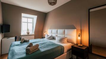 Una cama bien tendida con una colcha verde y almohadas blancas, una mesita de noche con una lámpara, una ventana con una planta y un televisor sobre un mueble.