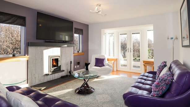 A modern living room with a fireplace, leather sofas, a glass coffee table, and a large flat-screen TV mounted on the wall. - 3 Croftside (Aviemore)
