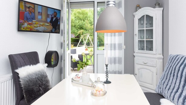 Zona pranzo con tavolo, sedie, lampada a sospensione, mobile contenitore e televisore appeso al muro.