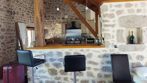 A rustic kitchen with a stone wall, wooden beams, and a central stove. There are two bar stools and a suitcase in the foreground. - Large house in the Morvan (Lormes)