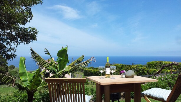 A wooden table with two chairs, a bottle of wine, and glasses on a grassy area with a view of the ocean.
