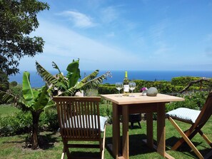 Una mesa de madera con dos sillas, una botella de vino y copas sobre una zona cubierta de césped con vista al océano.