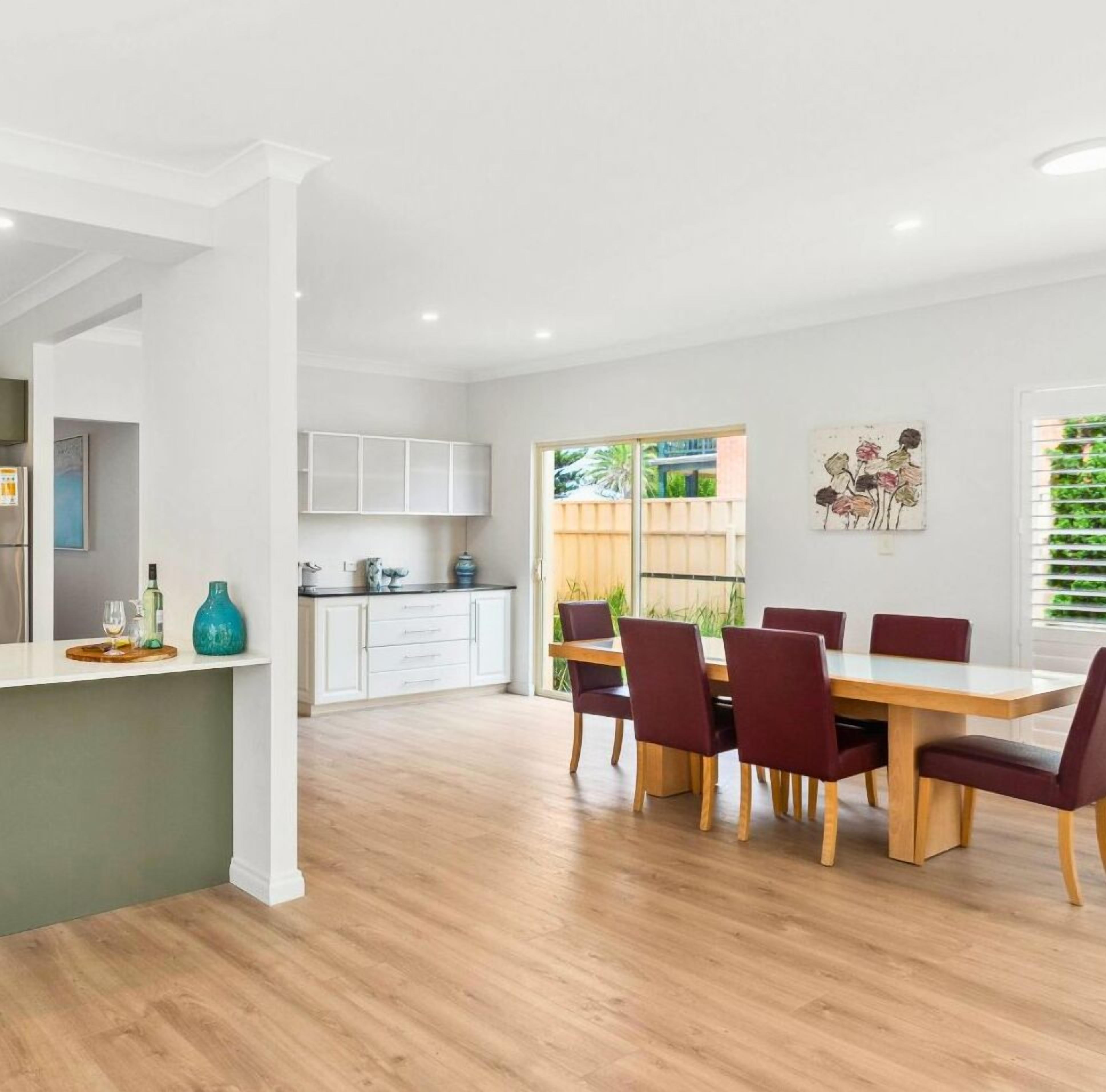 A modern kitchen with white cabinets, a dining area with a wooden table and red chairs, and a large window with a view of greenery.
