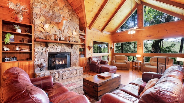 A cozy cabin interior with a stone fireplace, leather sofas, and a large window overlooking the forest. - Hazy Hideaway - Cherry Log, GA (Cherry Log)