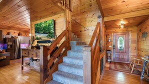 A wooden cabin interior with a staircase, kitchen, and a large window overlooking a forest. - Hazy Hideaway - Cherry Log, GA (Cherry Log)