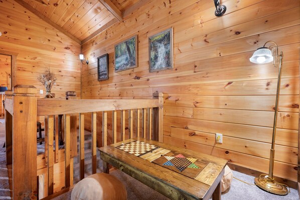 Interior de uma cabana de madeira com um banco rústico, uma mesa coberta por um tecido xadrez e um abajur.