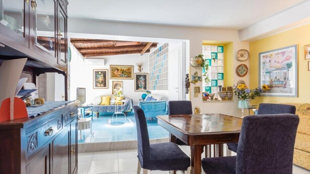 A spacious living area with a dining table, blue chairs, and a blue sofa. There is a kitchen area with cabinets and a refrigerator.