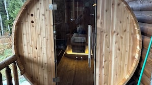 A wooden sauna with a glass door, a wooden deck, and a view of trees outside.