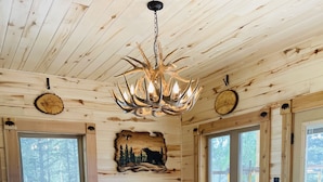 A large wooden dining table with a chandelier, surrounded by wooden chairs, in a cabin with wooden walls and windows.