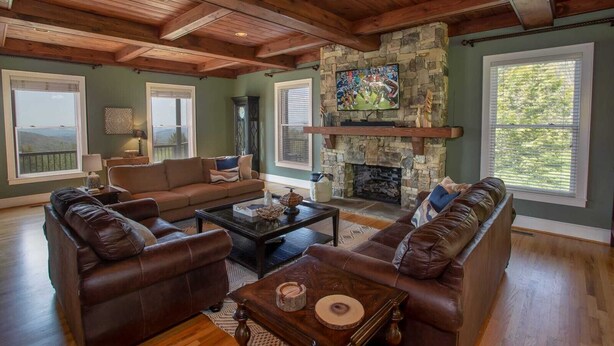 A spacious living room with a stone fireplace, leather sofas, and large windows offering a view of the outdoors. - Spectacular Estate with Breathtaking Views (Banner Elk)