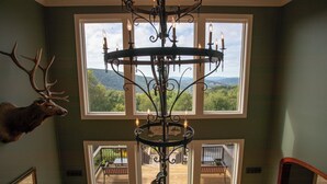 A room with a chandelier, a mounted deer head, and a view of a deck through large windows. - Spectacular Estate with Breathtaking Views (Banner Elk)