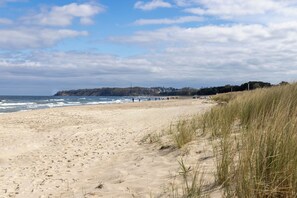 Une plage de sable parsemée d’empreintes, une dune herbeuse et, au loin, une côte bordée de bâtiments.