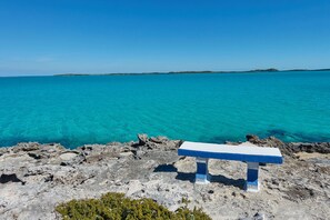 A bench on a rocky shoreline with clear turquoise water and distant land. - Ocean Front Villa -Crab House- (George Town)