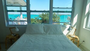 Ein Bett mit weißer Bettwäsche, zwei Nachttische mit Lampen und Blick auf das türkisfarbene Meer durch große Fenster.