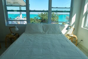 A bed with white bedding, two bedside lamps, and a view of a turquoise sea through large windows. - Ocean Front Villa -Crab House- (George Town)