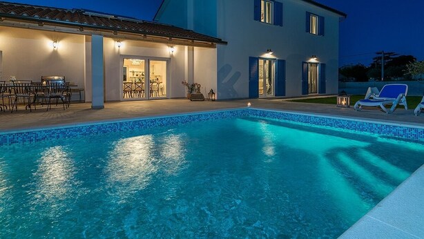 Una piscina con borde de azulejos, un patio con mesa y sillas, y un edificio con tejado y contraventanas azules.