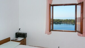 A bedroom with a bed, a bedside table with a radio, and a window with a view of a lake and trees.