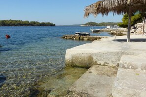 A clear waterfront with boats, a thatched-roof structure, and a rocky shoreline.