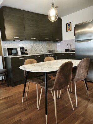Eine moderne Küche mit dunklen Schränken, einem Edelstahl-Kühlschrank, einem Esstisch mit Marmorplatte und vier Stühlen sowie einer Pendelleuchte.