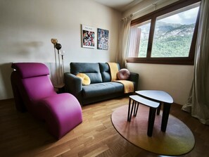 A living room with a sofa, a chair, a small table, and a window with a view of a mountainous landscape.