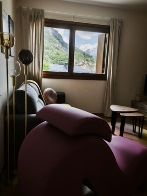 Ein modernes Wohnzimmer mit einer Couch, einem Tisch und Blick auf eine gebirgige Landschaft durch ein großes Fenster.
