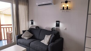 Una sala de estar moderna con un sofá gris, cojines estampados, una mesa de centro blanca y un ventanal corredero que da a un balcón exterior.