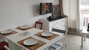 Comedor con una mesa blanca para cuatro, un televisor montado en la pared y vistas al exterior a través de una ventana con cortinas.