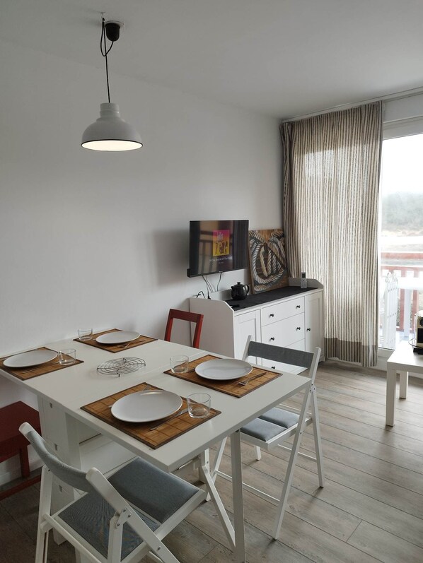 A dining area with a white table set for four, a television mounted on the wall, and a view of the outdoors through a window with curtains.