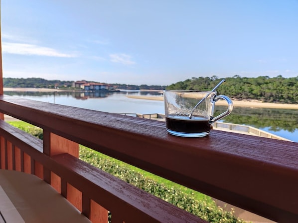 A glass mug with a spoon on a wooden railing overlooking a calm lake with buildings in the background.
