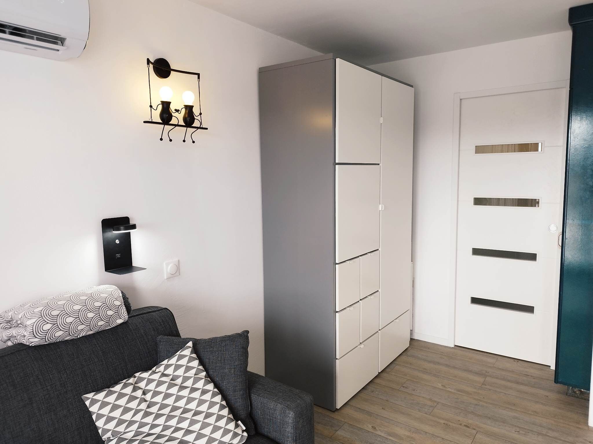 Une pièce moderne avec un canapé gris, des coussins à motifs, un luminaire fixé au mur, une armoire et une porte.