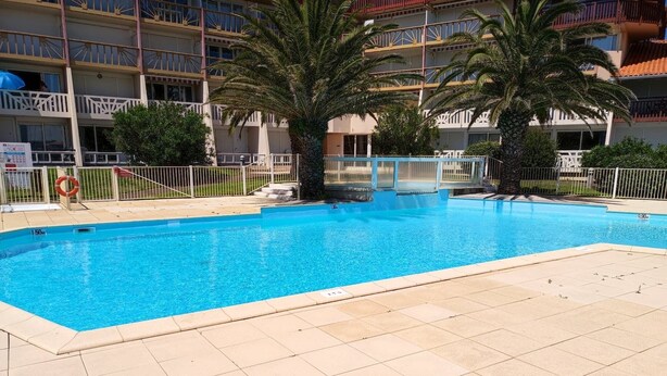 Una piscina con borde de baldosas, rodeada de palmeras y un edificio de varios pisos con balcones.