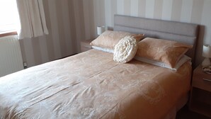 Ein ordentlich bezogenes Bett mit einer blumigen Tagesdecke und zwei Kissen, ein Nachttisch mit Lampe und Tasse, und ein Fenster mit Vorhängen.
