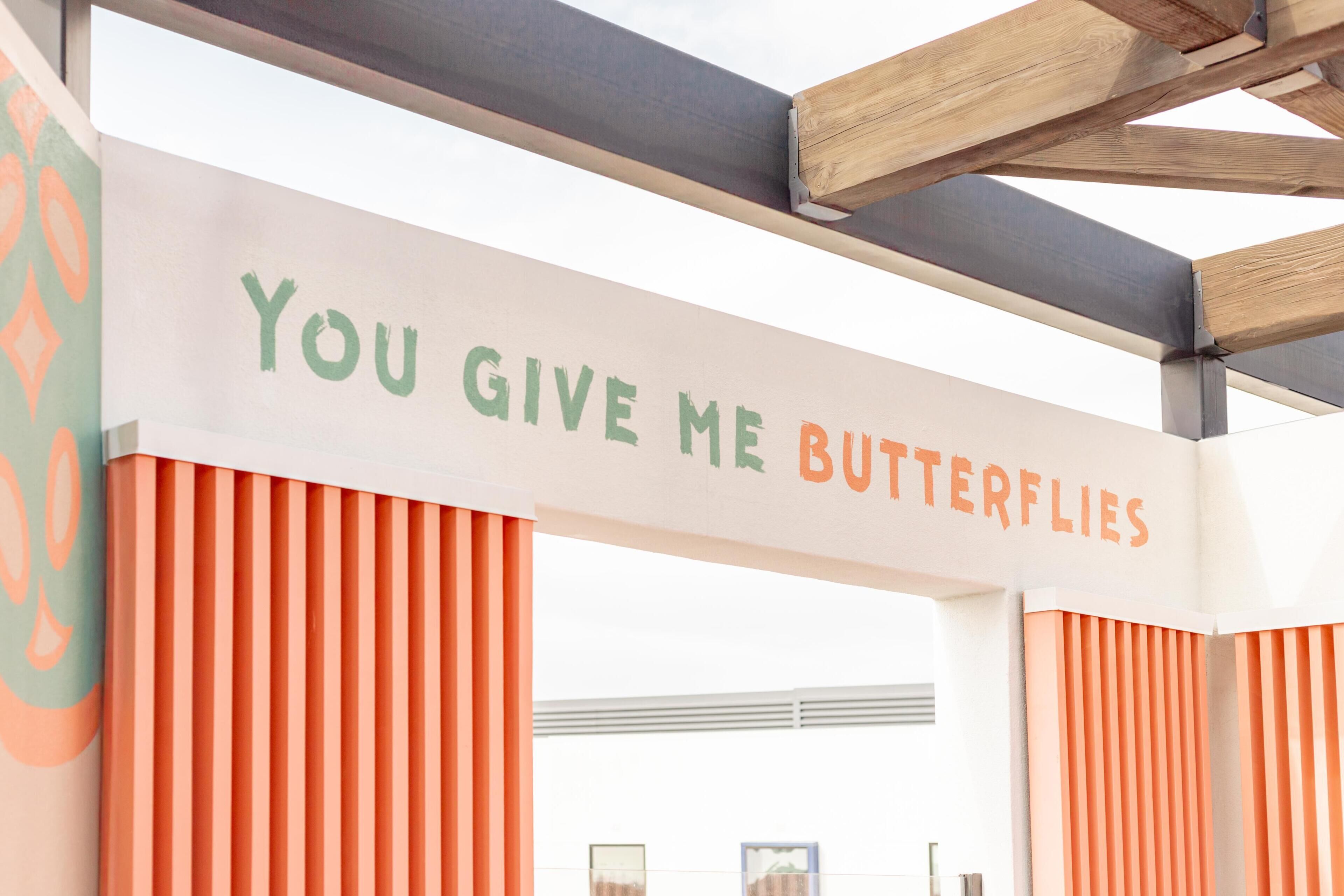A sign with the text 'YOU GIVE ME BUTTERFLIES' in a building with orange vertical panels and a wooden structure overhead.